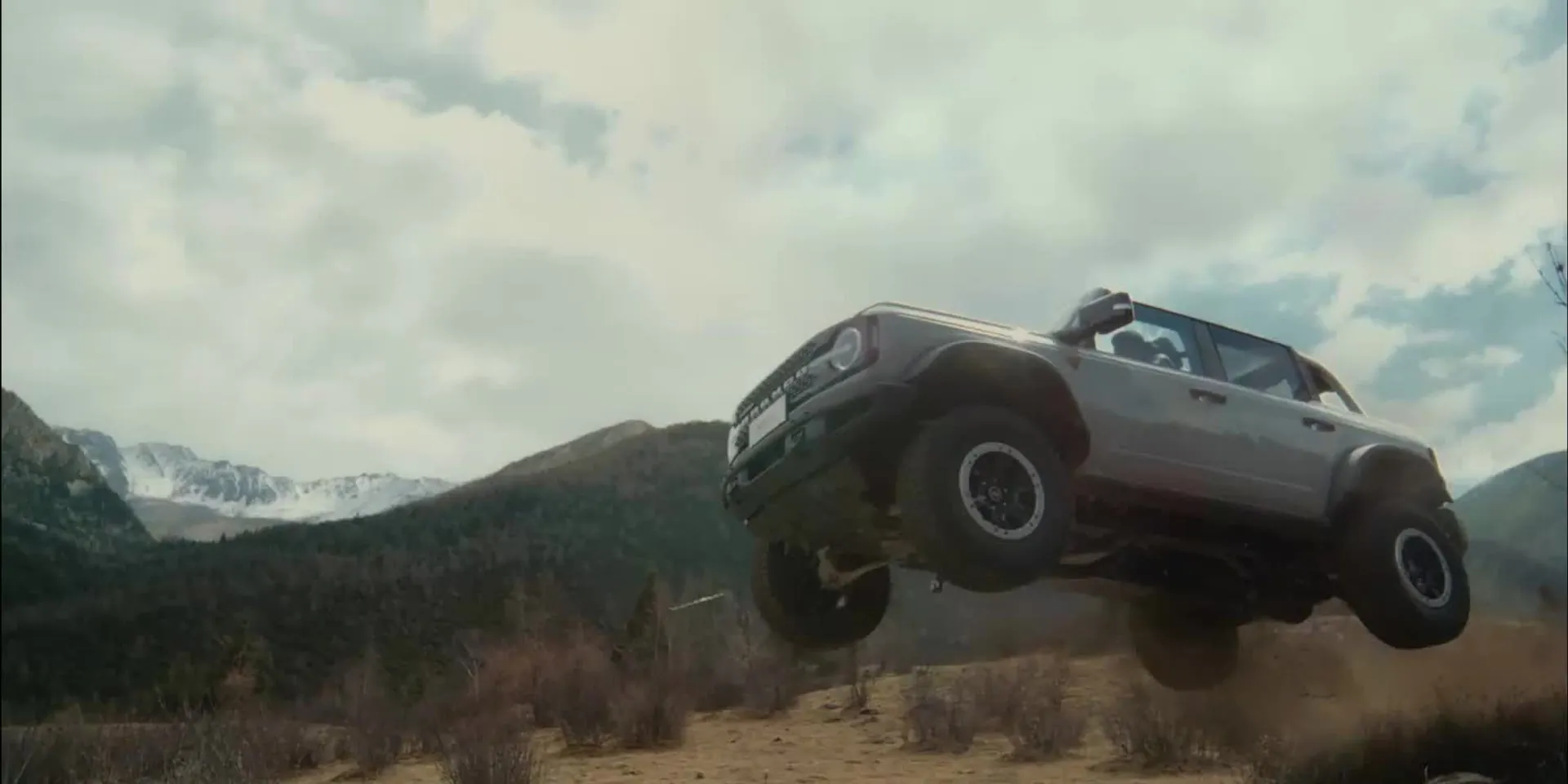 A Ford Bronco in mid-air over rough ground, mountains behind